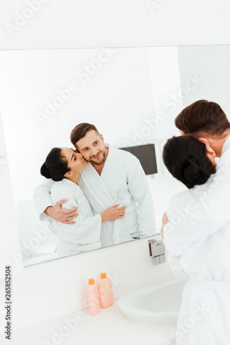 selective focus of happy woman hugging and kissing cheek of man while looking at mirror