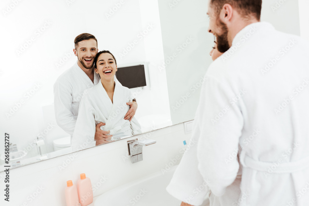 selective focus of bearded man looking at mirror with cheerful brunette woman while hugging in bathroom selective focus of bearded man looking at mirror with cheerful brunette woman while hugging in bathroom
