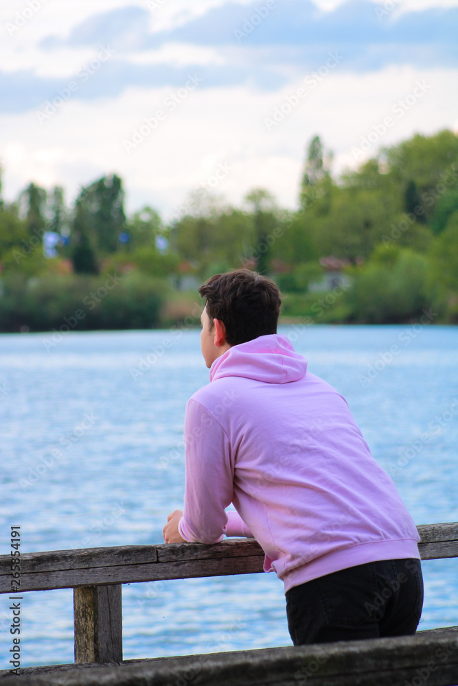 Young man contemplating a lake.  Teenager with a pink sweater who is standing upright against a railing of a bridge. 