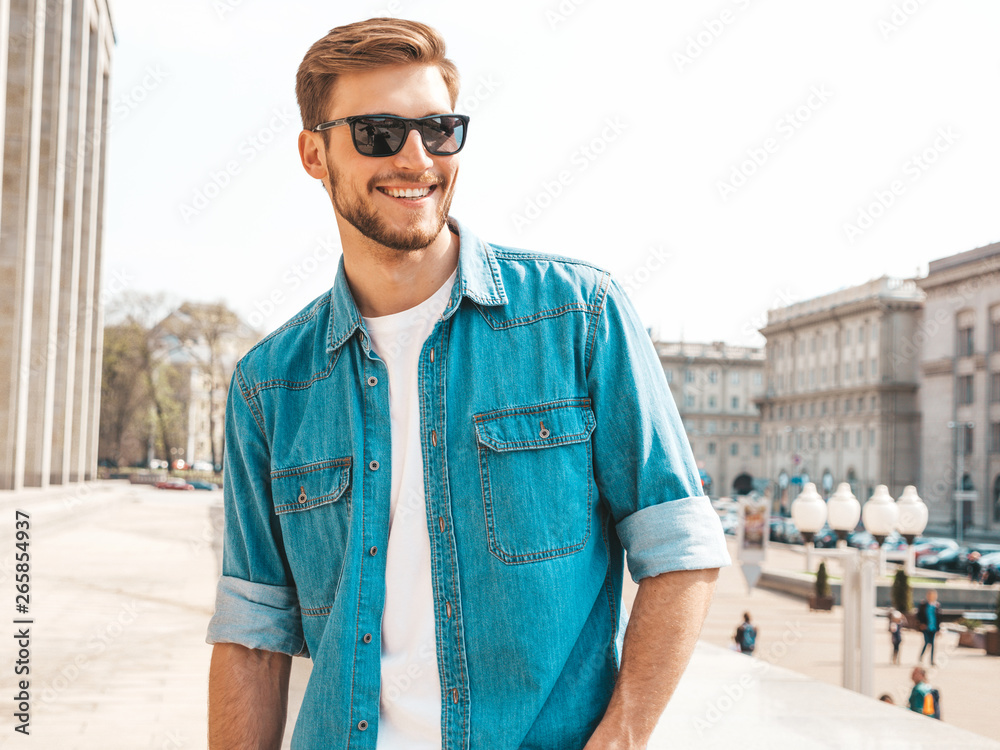 Portrait of handsome smiling stylish hipster lumbersexual businessman model. Man dressed in jeans jacket clothes. Fashion male posing on the street background in sunglasses Portrait of handsome smiling stylish hipster lumbersexual businessman model. Man dressed in jeans jacket clothes. Fashion male posing on the street background in sunglasses