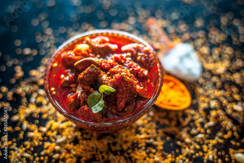 Popular Indian & Asian pickle of mango in a glass bowl i.e. Aam ka achar in a glass bowl or keri ka achar with its entire raw ingredients including spices and mango on black colored shiny surface.
