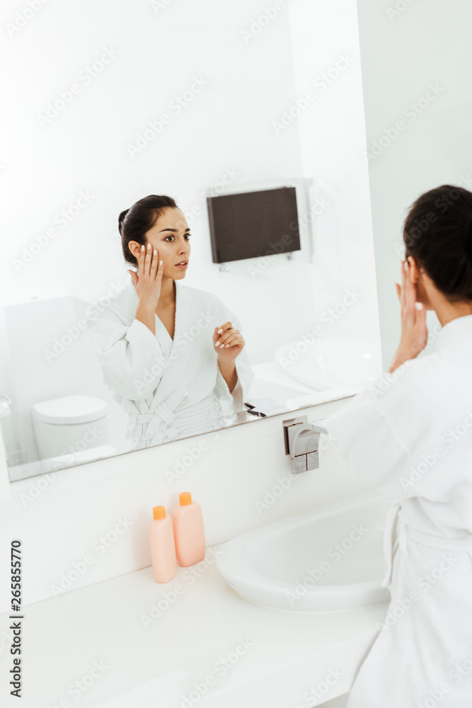selective focus of attractive brunette woman touching face while looking at mirror in bathroom