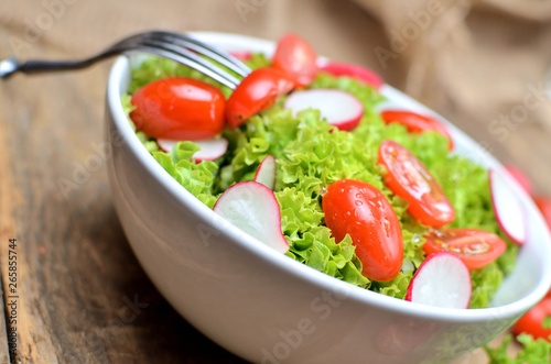 Close-up of green salad Lollo Biondo with tomatoes and radishes in a white bowl with fork on wooden table