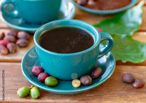 Cup with black coffee served outside with raw green, mature red and roasted coffee beans, decorated with green leaves from coffee plant