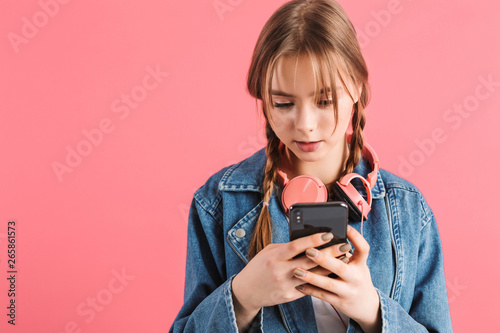 Young pretty girl with two braids in denim jacket with headphones dreamily using cellphone over pink background isolated