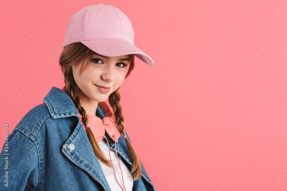 Portrait of young pretty joyful girl with two braids in denim jacket and cap with headphones on neck happily looking in camera over pink background isolated Portrait of young pretty joyful girl with two braids in denim jacket and cap with headphones on neck happily looking in camera over pink background isolated