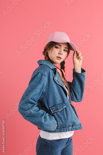 Young attractive girl with two braids in denim jacket and cap with headphones on neck holding hand in pocket dreamly looking in camera over pink background isolated © Anton