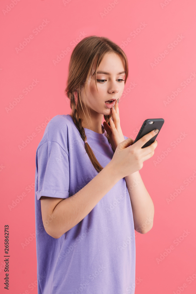 Young attractive girl with two braids in lilac t shirt amazedly using cellphone while spending time over pink background Young attractive girl with two braids in lilac t shirt amazedly using cellphone while spending time over pink background
