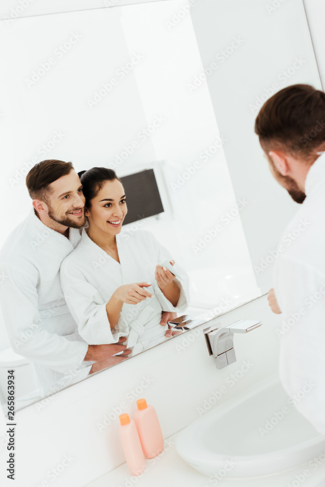 selective focus of happy man hugging attractive brunette woman holding toothpaste near mirror selective focus of happy man hugging attractive brunette woman holding toothpaste near mirror
