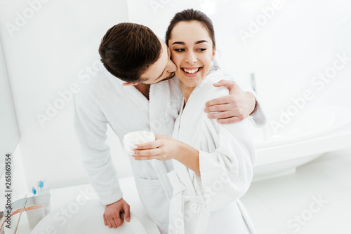 cheerful boyfriend kissing cheek of happy girl holding container with face cream