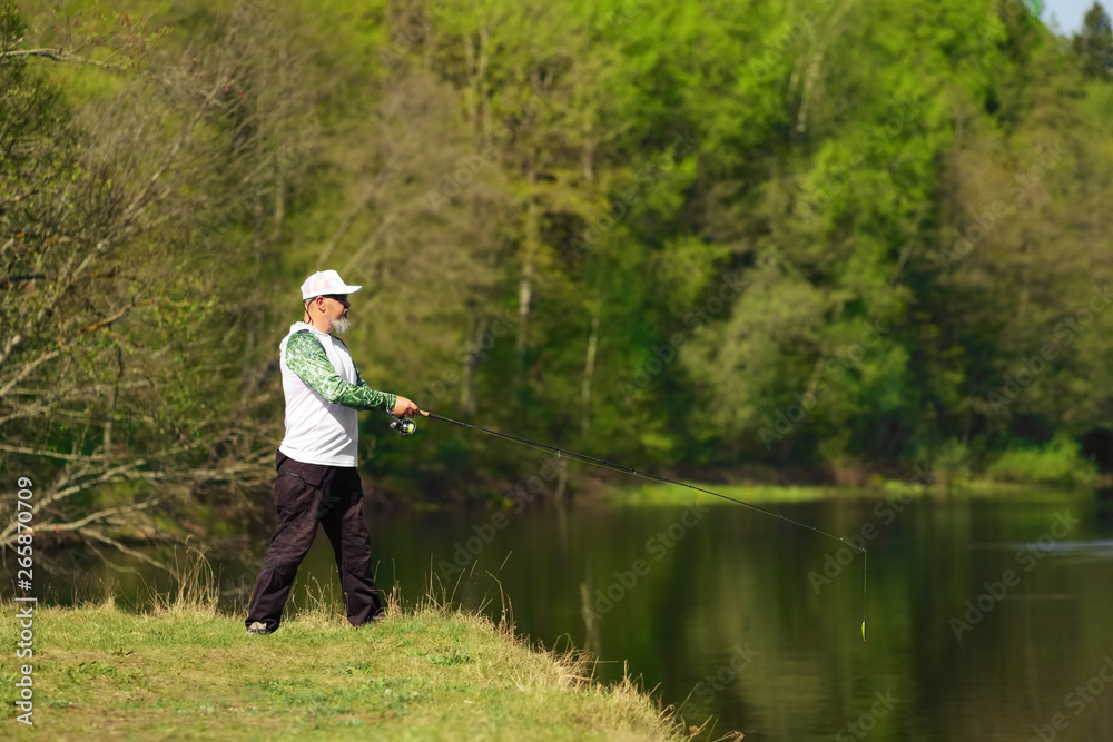 Fisherman with a spinning rod catching fish on a river at sunny summer day with green trees at background. Outdoor weekend activity. Photo with shallow depth of field taken at wide open aperture. Fisherman with a spinning rod catching fish on a river at sunny summer day with green trees at background. Outdoor weekend activity. Photo with shallow depth of field taken at wide open aperture.