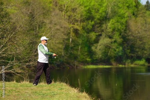 Fisherman with a spinning rod catching fish on a river at sunny summer day with green trees at background. Outdoor weekend activity. Photo with shallow depth of field taken at wide open aperture. © boomeart