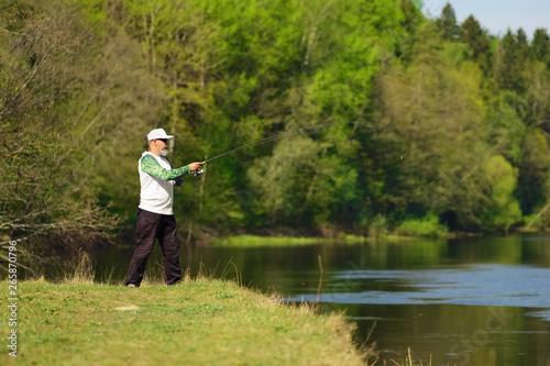Fisherman with a spinning rod catching fish on a river at sunny summer day with green trees at background. Outdoor weekend activity. Photo with shallow depth of field taken at wide open aperture.