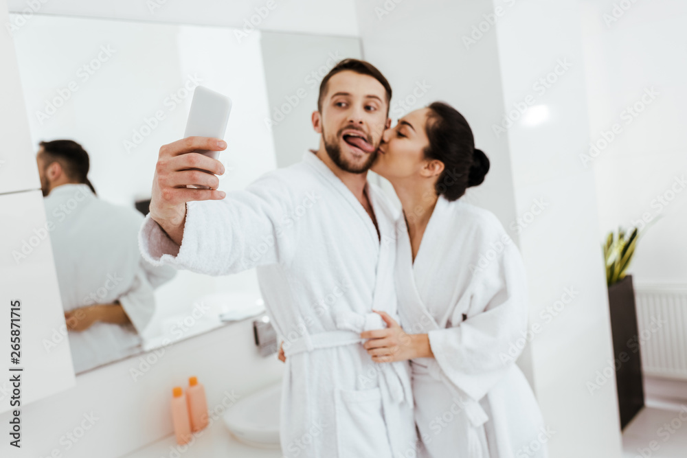 girlfriend kissing cheek of cheerful man showing tongue while taking selfie in bathroom girlfriend kissing cheek of cheerful man showing tongue while taking selfie in bathroom