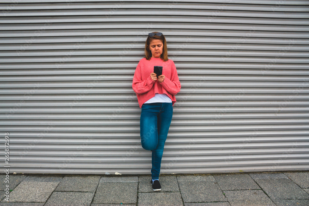 Blonde young woman in jeans and a big pink blouse using and looking at the smartphone – Girl holding and texting on her cellphone – Concept for teens with gadgets as a lifestyle Blonde young woman in jeans and a big pink blouse using and looking at the smartphone – Girl holding and texting on her cellphone – Concept for teens with gadgets as a lifestyle