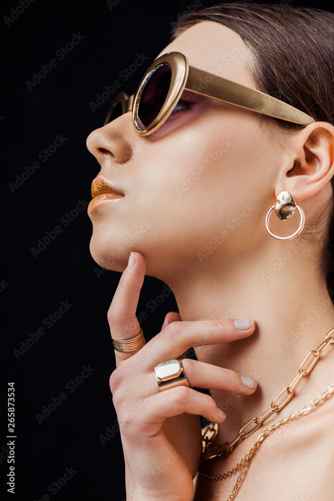 profile of young naked woman in sunglasses, golden earring, rings and necklaces isolated on black