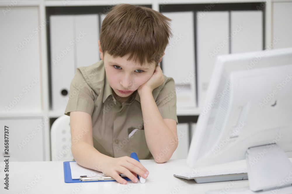 Sad schoolboy does homework. Upset boy behind desk with pen and notebook.The little boy thinks over the task Sad schoolboy does homework. Upset boy behind desk with pen and notebook.The little boy thinks over the task
