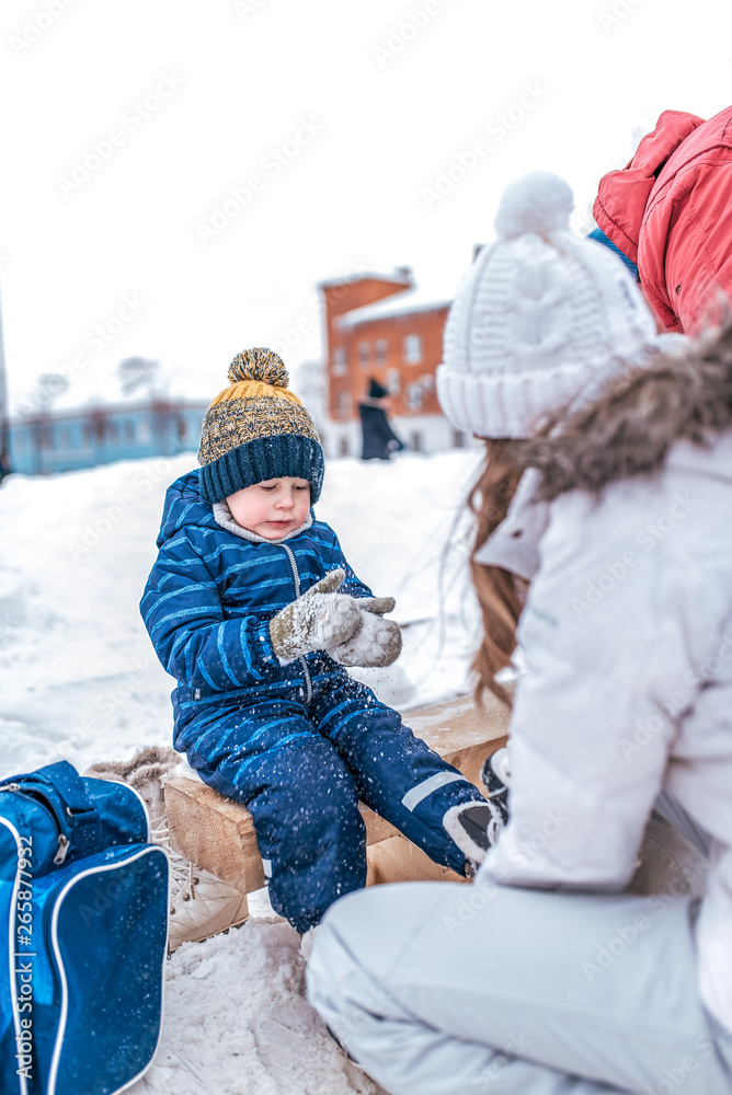 Young mother puts on skates in the winter on a public skating rink. Little boy in a blue jumpsuit, mittens in the snow. Learning ice skating in winter on the ice.