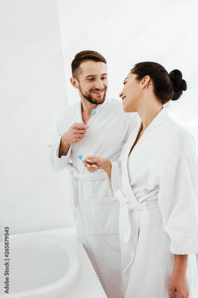 happy man holding toothbrush and looking at brunette woman in bathroom happy man holding toothbrush and looking at brunette woman in bathroom