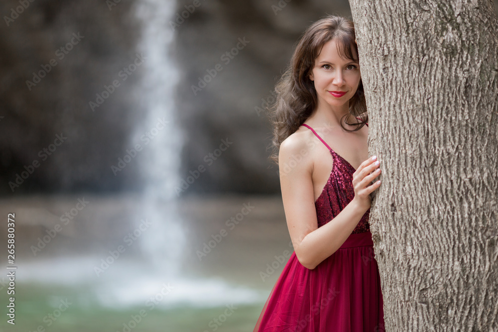 Young beautiful woman is wearing fashion red dress walking in forest near the waterfall
