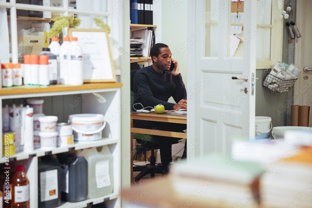 Confident young male owner talking on smart phone while sitting at desk in furniture store Confident young male owner talking on smart phone while sitting at desk in furniture store