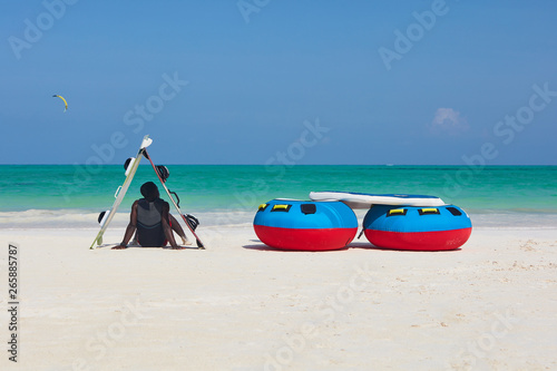 Vertical back view of a man in shade of boards sitting down facing the ocean.