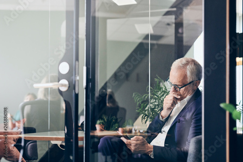 Mature male manager using smart phone seen through glass wall at creative office