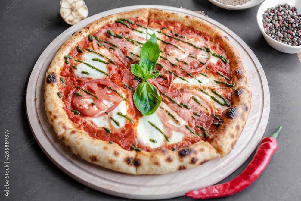 Pizza Margherita with green salsa and leaf of basyl on wooden plate, garlic, red pepper and bowl with spices near. Black background, top view, flat lay, vertical image. Natural light. Pizza Margherita with green salsa and leaf of basyl on wooden plate, garlic, red pepper and bowl with spices near. Black background, top view, flat lay, vertical image. Natural light.