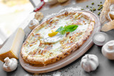 Pizza with ham, cheese and fried egg in the middle on the wooden plate. Garlic, champignons, spices near on the table, piese of cheese. Horizontal. Natural light. Black background. Shallow focus.