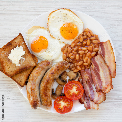 Full English breakfast with fried eggs, sausages, beans, bacon and toasts on a white wooden background. Top view, flat lay. From above, overhead.