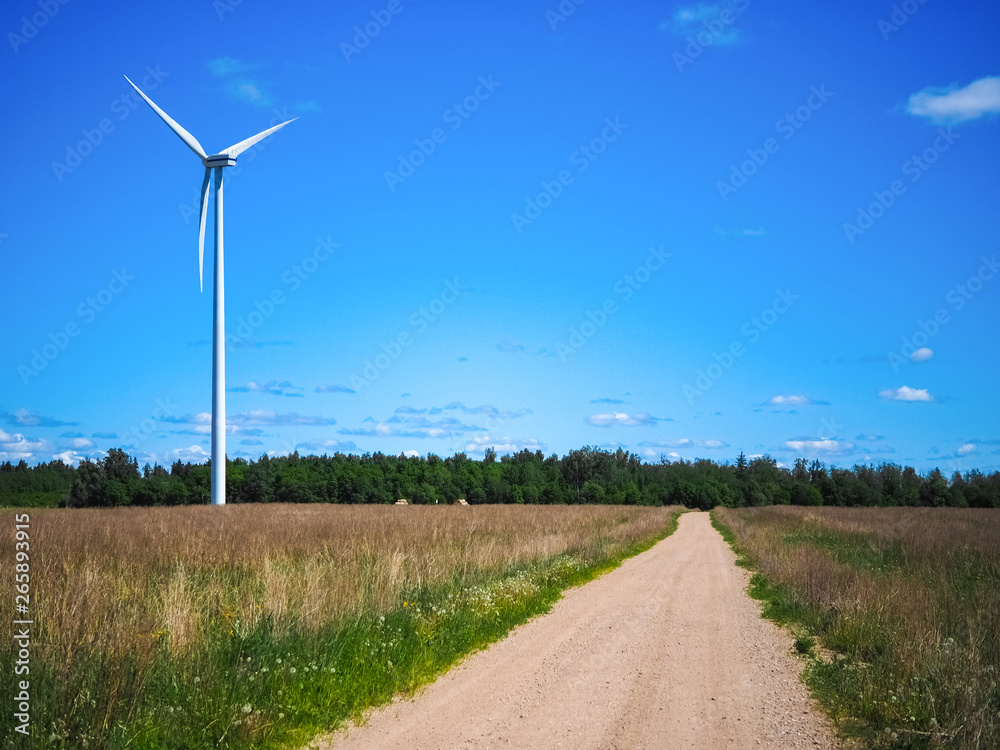 Wind power generator on an empty field. Green forest in the background. The road leads to the wind turbine. Green energy on a sunny day. Blue sky with clouds. Ecology and energy conservation concept.