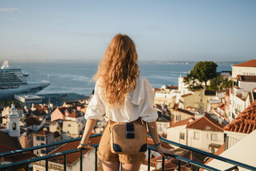 Blonde woman standing on the balcony and looking at coast view of the southern european city with sea during the sunset, wearing hat, cork bag, safari shorts and white shirt