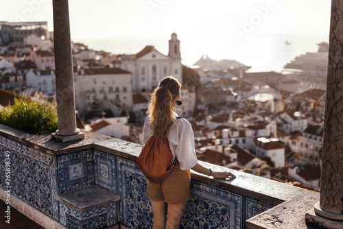 Blonde woman standing on the balcony and looking at coast view of the southern european city with sea during the sunset, wearing hat, cork bag, safari shorts and white shirt