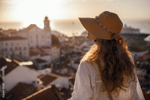 Blonde woman standing on the balcony and looking at coast view of the southern european city with sea during the sunset, wearing hat, cork bag, safari shorts and white shirt