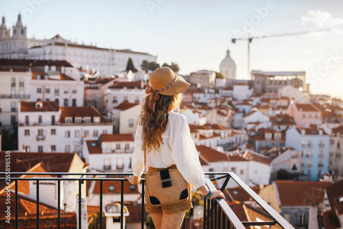 Blonde woman standing on the balcony and looking at coast view of the southern european city with sea during the sunset, wearing hat, cork bag, safari shorts and white shirt
