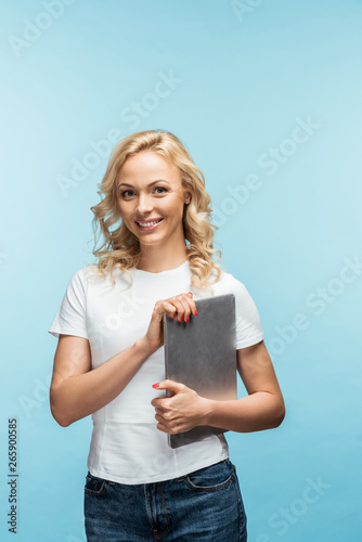happy blonde woman holding laptop and looking at camera on blue
