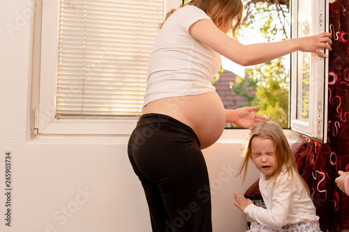 Young pregnant woman standing by the window, arguing to her little daughter crying discipline conflict family violence
