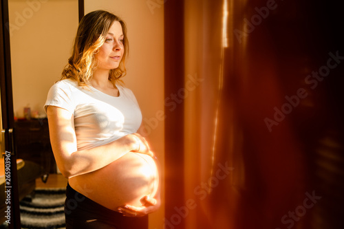 Young beautiful pregnant woman standing by the window looking holding her belly waiting for the baby