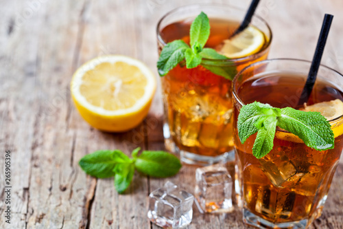 Cold iced tea with lemon, mint leaves and ice cubes in two glasses on rustic wooden table background.