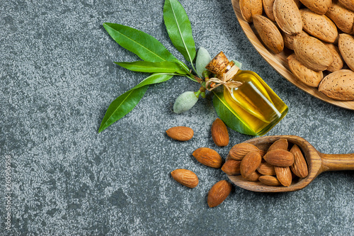Glass bottle of Almond oil and almond nuts in wooden shovel, almonds with shell in bamboo bowl on grey rustic table with green fresh raw almonds on almond tree branch