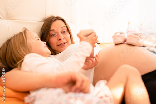 Pregnant mother and young daughter small child laying in bed having fun with smart phone looking family photos taking selfies