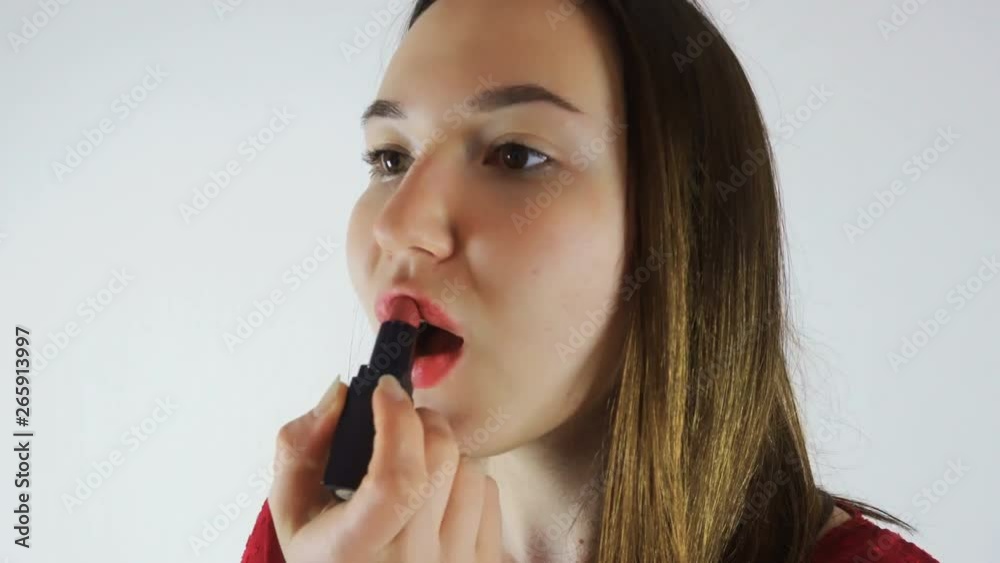 A young brunette woman in a red blouse applies make-up, paints her lips with lipstick. She looks in the mirror. Close-up A young brunette woman in a red blouse applies make-up, paints her lips with lipstick. She looks in the mirror. Close-up