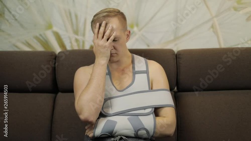 Painful, bored man with a broken arm wearing arm brace sitting on a sofa photo