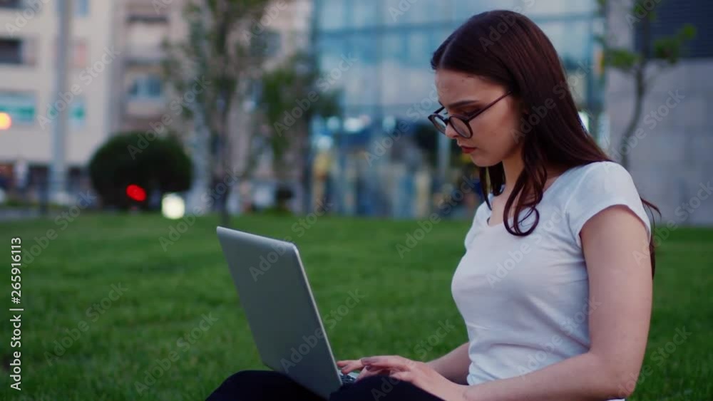 Young pretty trendy Caucasian woman typing keyboard while sitting on lawn, charmingly smiles. Girl using laptop outside. Positive emotions, good mood, happiness. Female portrait Young pretty trendy Caucasian woman typing keyboard while sitting on lawn, charmingly smiles. Girl using laptop outside. Positive emotions, good mood, happiness. Female portrait