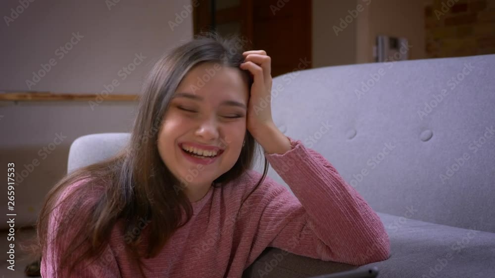 Closeup shoot of young pretty brunette caucasian female browsing on the tablet and laughing happily in a cozy apartment indoors Closeup shoot of young pretty brunette caucasian female browsing on the tablet and laughing happily in a cozy apartment indoors