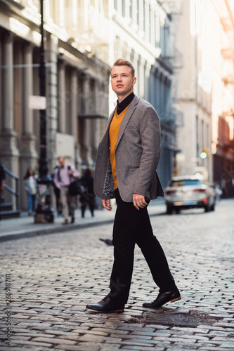 Handsome blonde man walking on city street wearing a smart casual outfit with a jacket and black pants in New York City SoHo district.