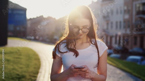 Joyful young woman in white t-shirt nerdy like glasses walks down the city and uses smartphone for texting, cutely smiling in bright sunlight. Social networks, messangers. Close up, sun lens photo