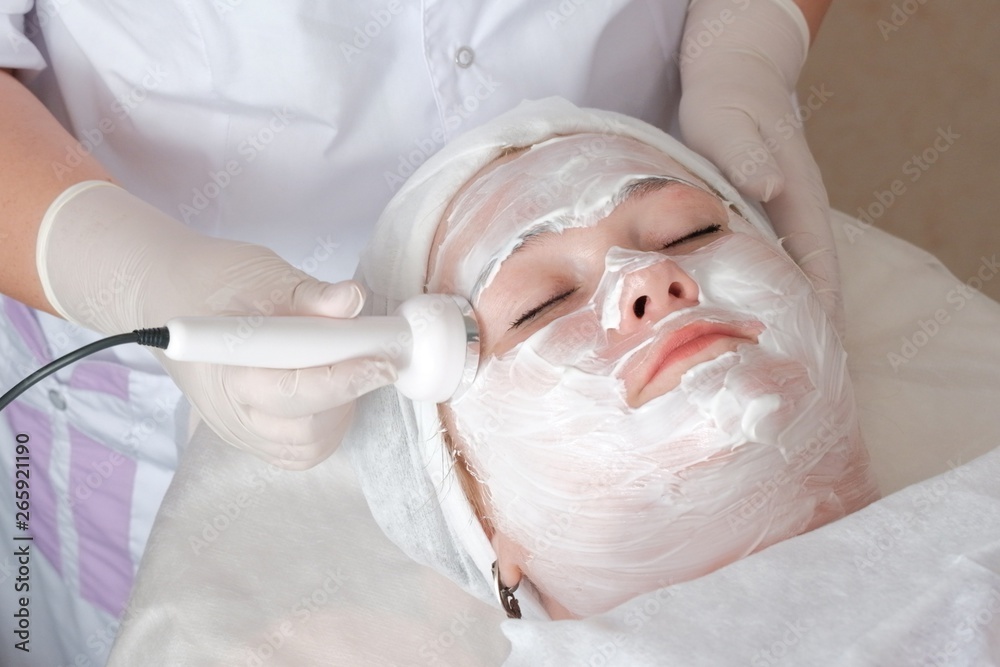 Fototapeta premium The beautician in white gloves holds the electrode of the massage apparatus and smoothes mimic wrinkles on the client's face. Anti-aging effect. Fat woman on the procedure in the cosmetology center.