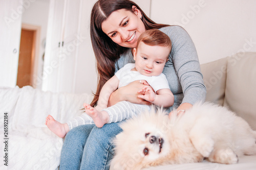 Young mother brunette woman with baby boy in arms play with dog Pomeranian Spitz at home