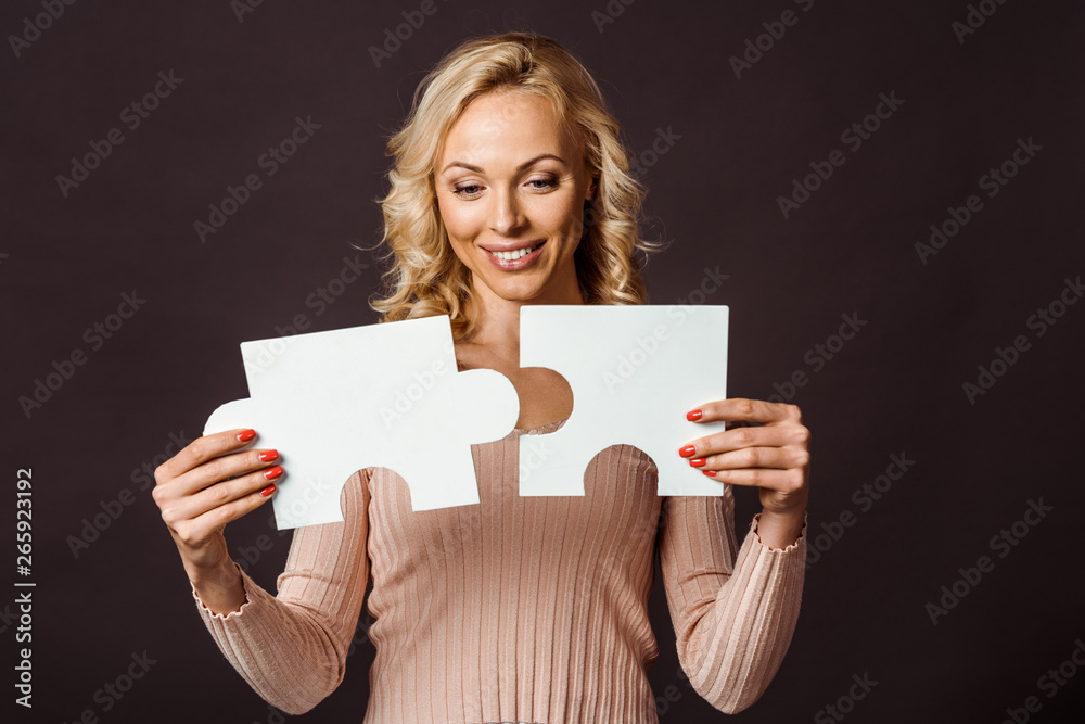 Naklejka premium happy blonde woman connecting jigsaw pieces isolated on black